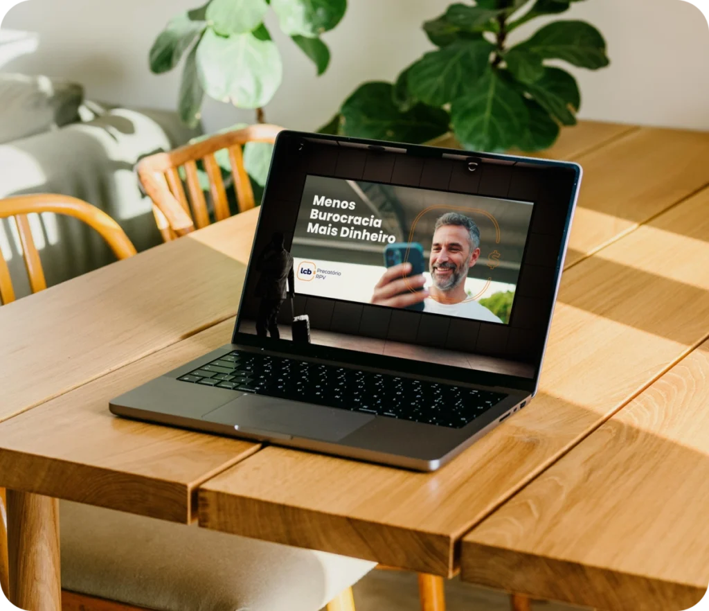 Laptop mostrando anúncio "Menos Burocracia Mais Dinheiro" da LCB Assessoria. Mesa de madeira, plantas ao fundo.