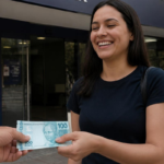 Mão entrega nota de 100 reais para mulher sorrindo em frente a banco