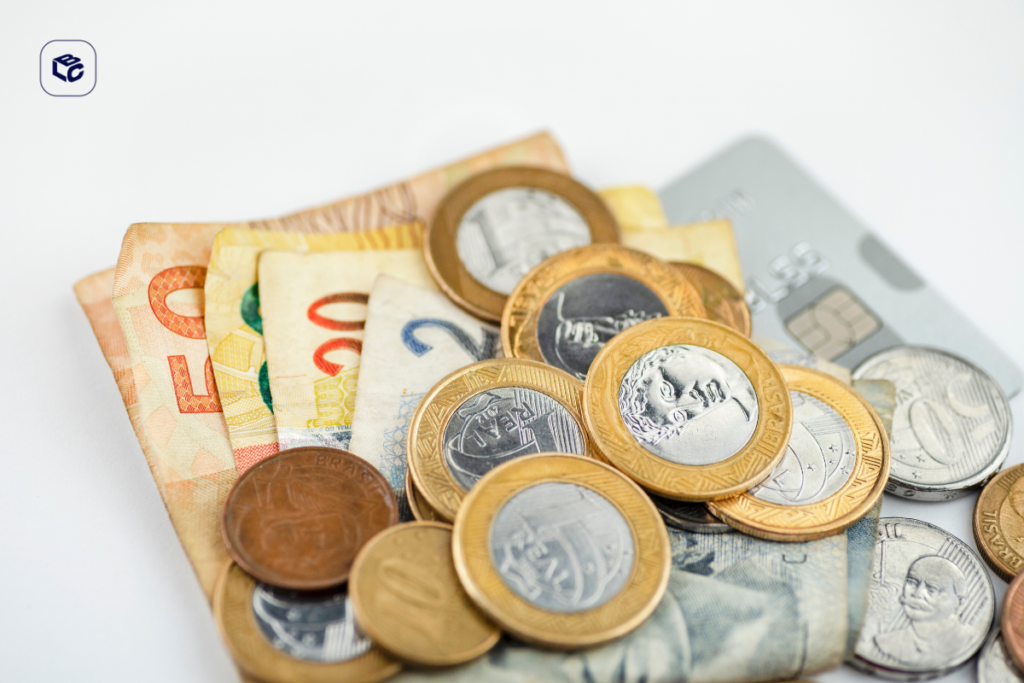 Cédulas e moedas de real espalhadas sobre uma superfície clara, ao lado de um cartão bancário parcialmente visível. A imagem representa dinheiro, finanças pessoais, pagamentos e recursos financeiros.