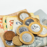 Cédulas e moedas de real espalhadas sobre uma superfície clara, ao lado de um cartão bancário parcialmente visível. A imagem representa dinheiro, finanças pessoais, pagamentos e recursos financeiros.